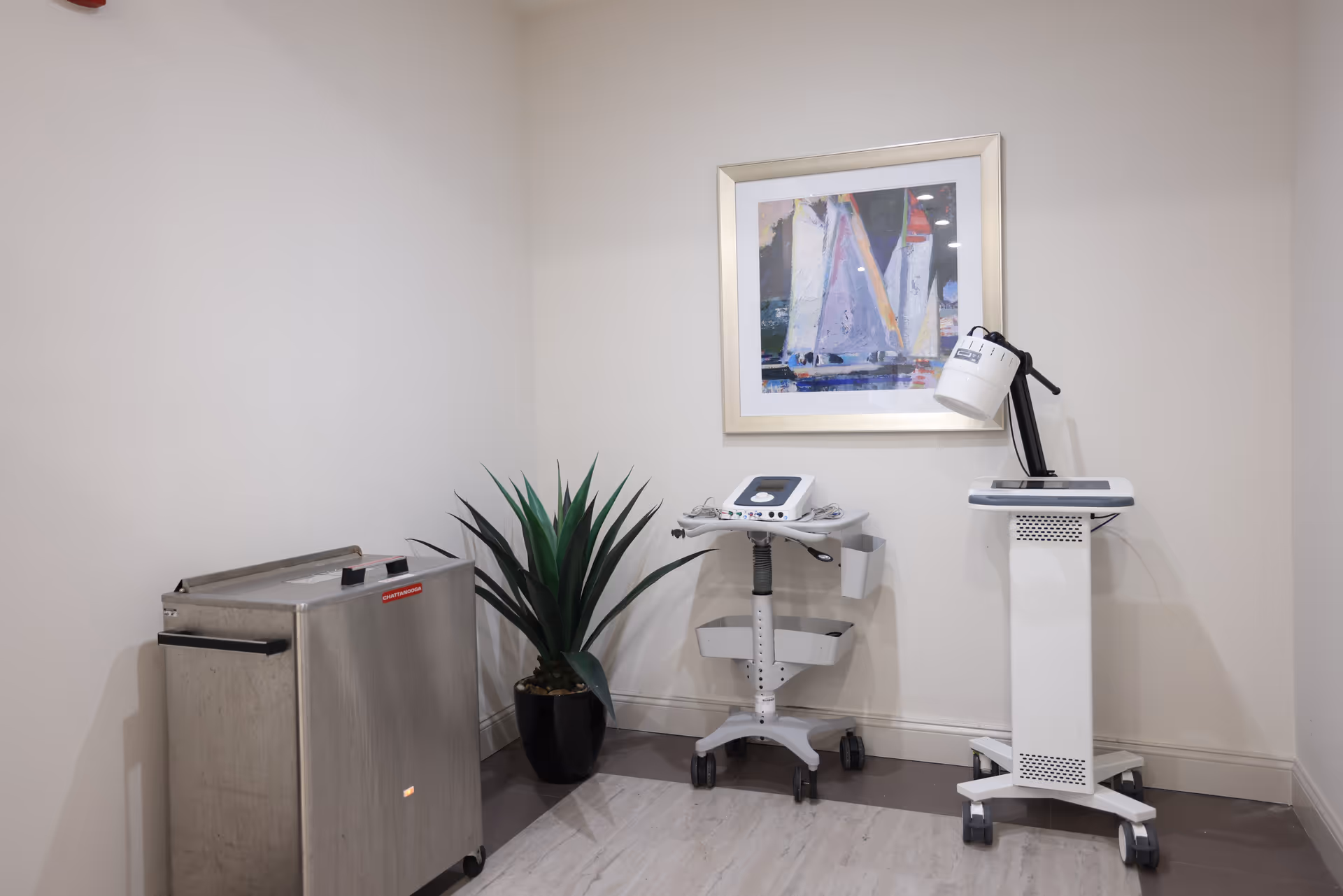 A compact medical examination room with wheeled equipment, a plant, and wall art.