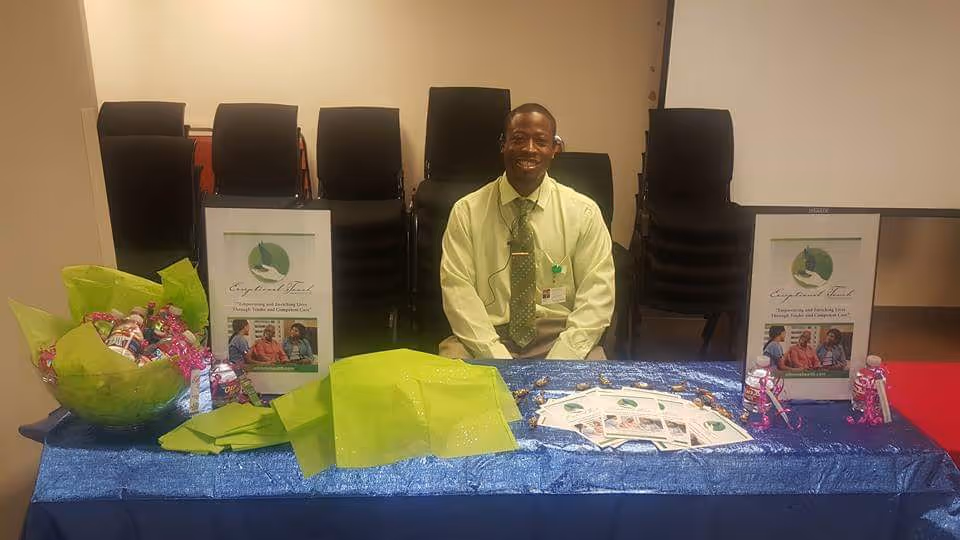 Smiling staff member sits behind a table with brochures and gift bags in a conference room.