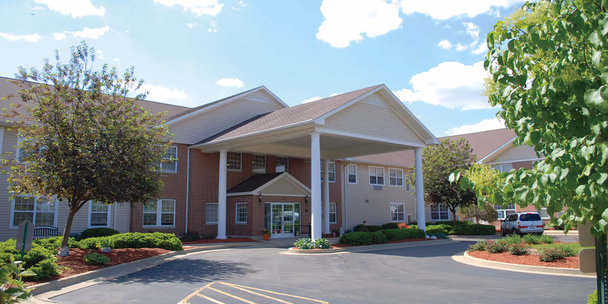 Exterior view of Bowman Estates of Danville, showing a two-story building with a covered entrance supported by white columns, surrounded by landscaped greenery and trees under a partly cloudy sky.