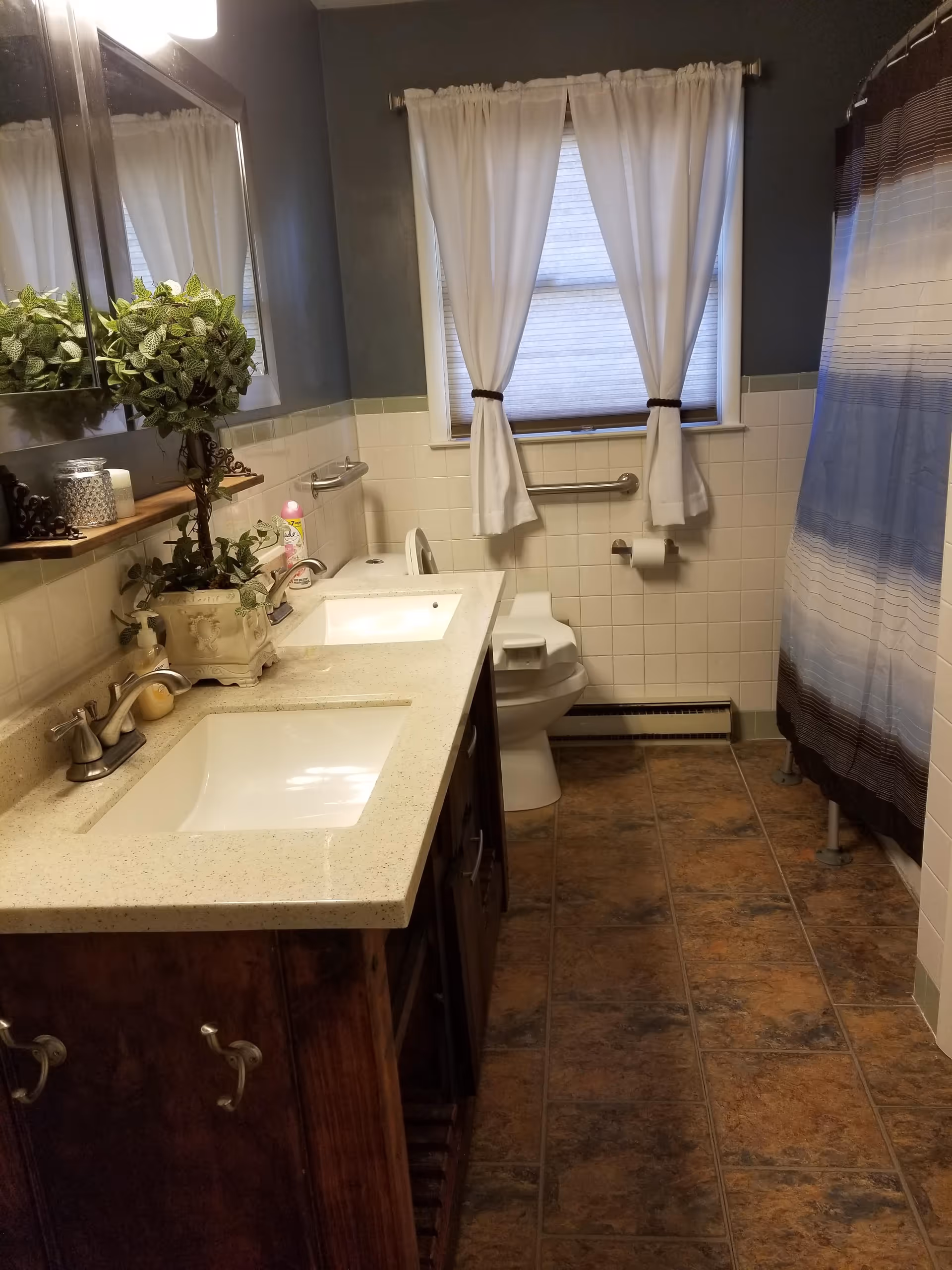 Bathroom with a double-sink vanity, mirror, toilet with a grab bar, and a shower with a striped curtain.