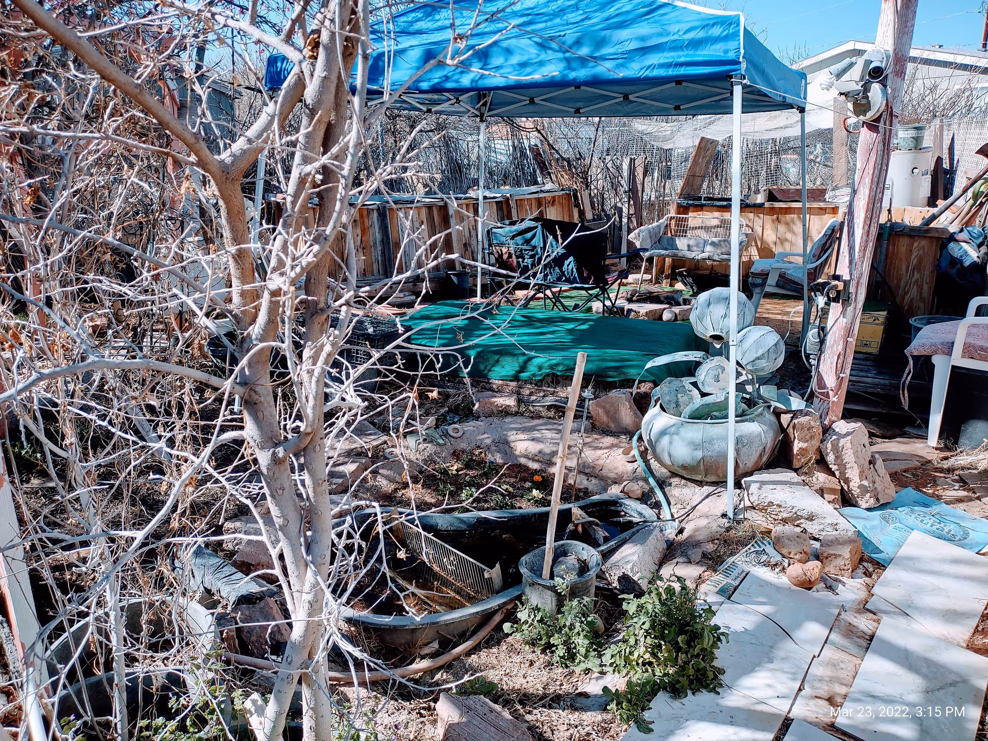 Uneven cluttered backyard with a blue canopy, chairs, rocks, and scattered debris.