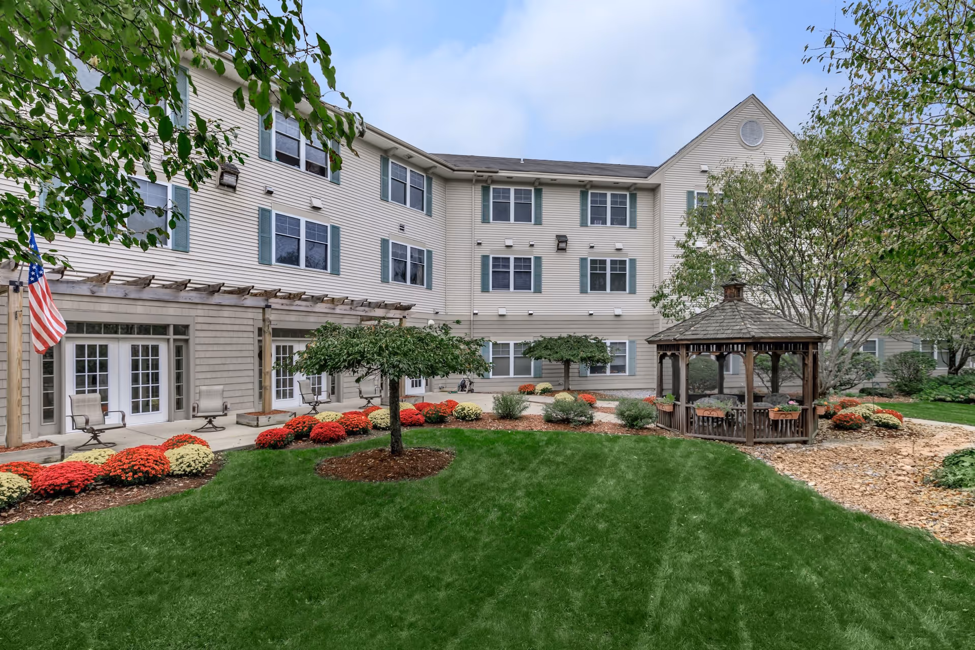 Outdoor courtyard area of a senior living facility with a well-maintained green lawn, colorful flower beds, small trees, a wooden gazebo, and a three-story beige building with multiple windows and green shutters in the background.