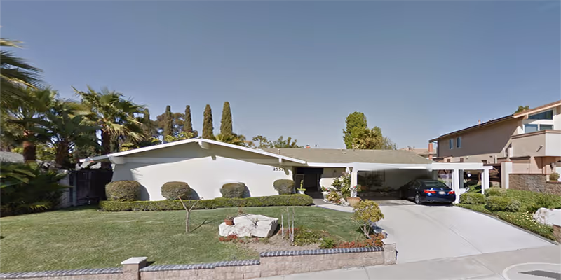 Single-story white senior living cottage with a driveway, carport, and manicured front yard.