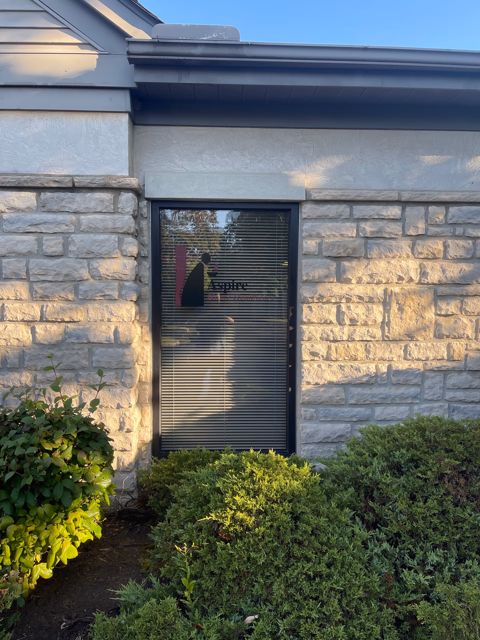 Stone building exterior with a front doorway, window blinds, Aspire logo, and landscaped shrubs.