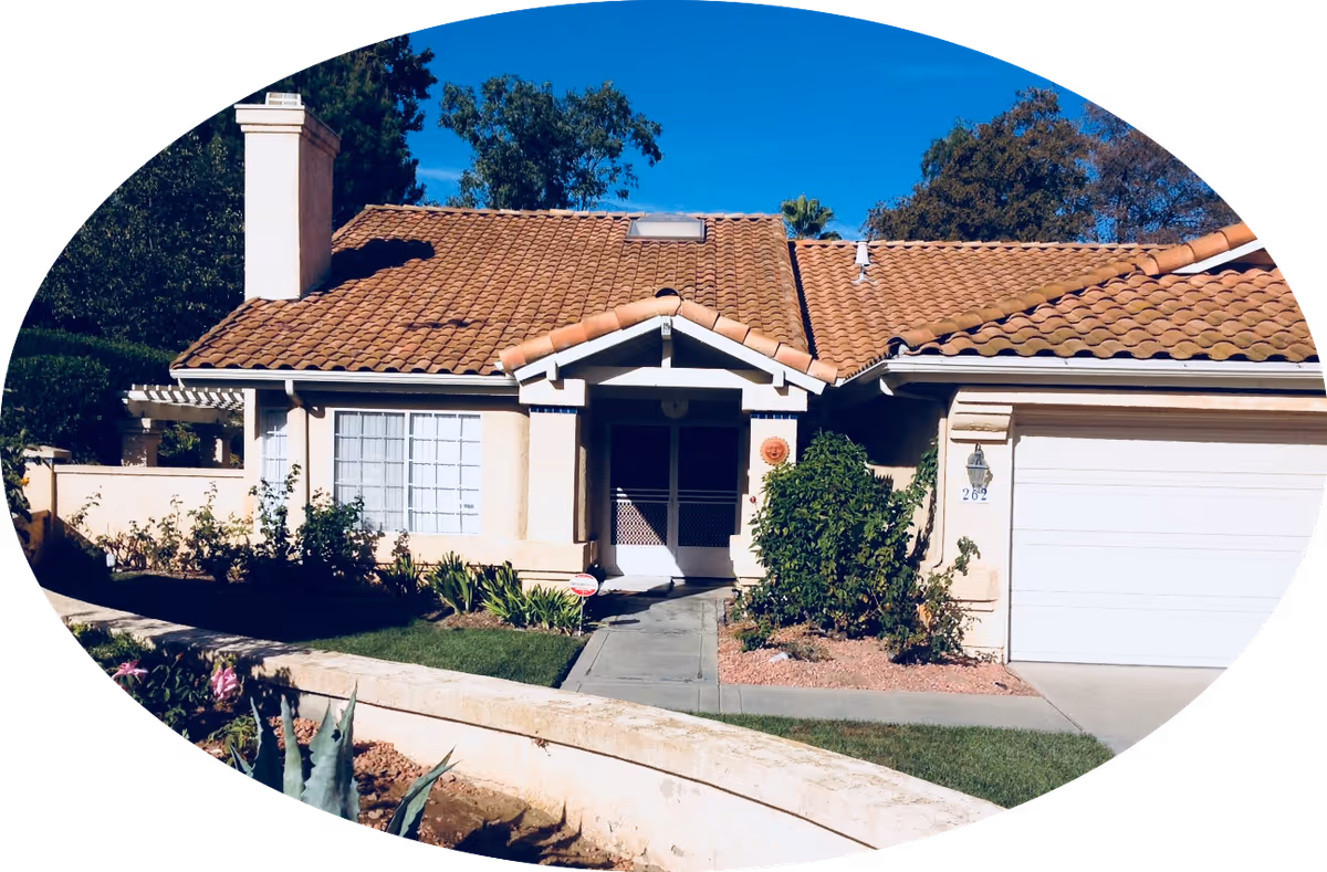 Front exterior view of a single-story house with a tiled roof, a chimney, a garage door on the right, and a walkway leading to the front door. The house is surrounded by a small garden with bushes and plants, and there is a clear blue sky in the background.