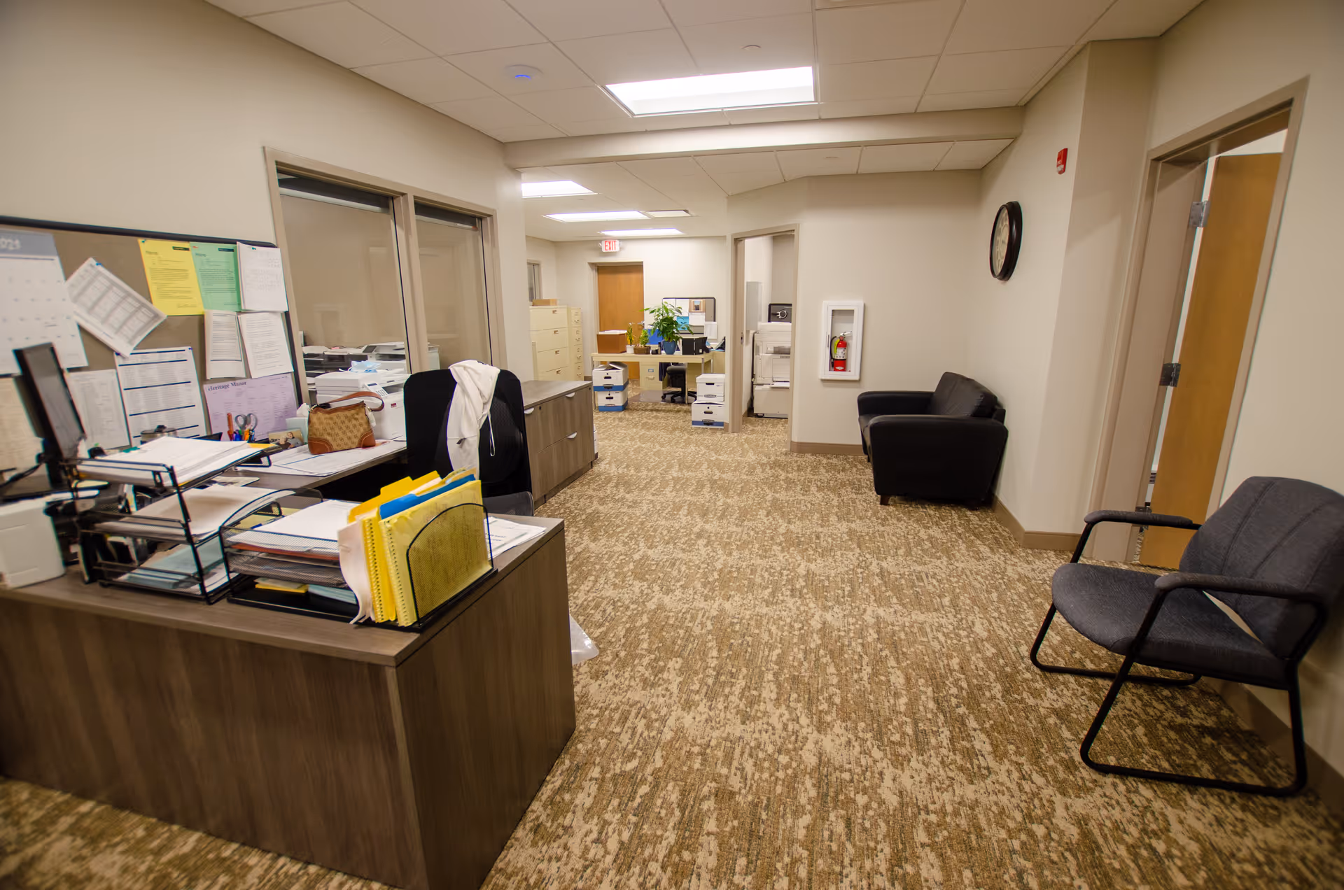 Interior view of an office area in a senior living facility with a desk covered in paperwork and office supplies on the left, two chairs on the right side, a black couch against the far wall, filing cabinets, and a clock on the wall. The space is well-lit with overhead fluorescent lights and has beige walls and patterned carpet flooring.
