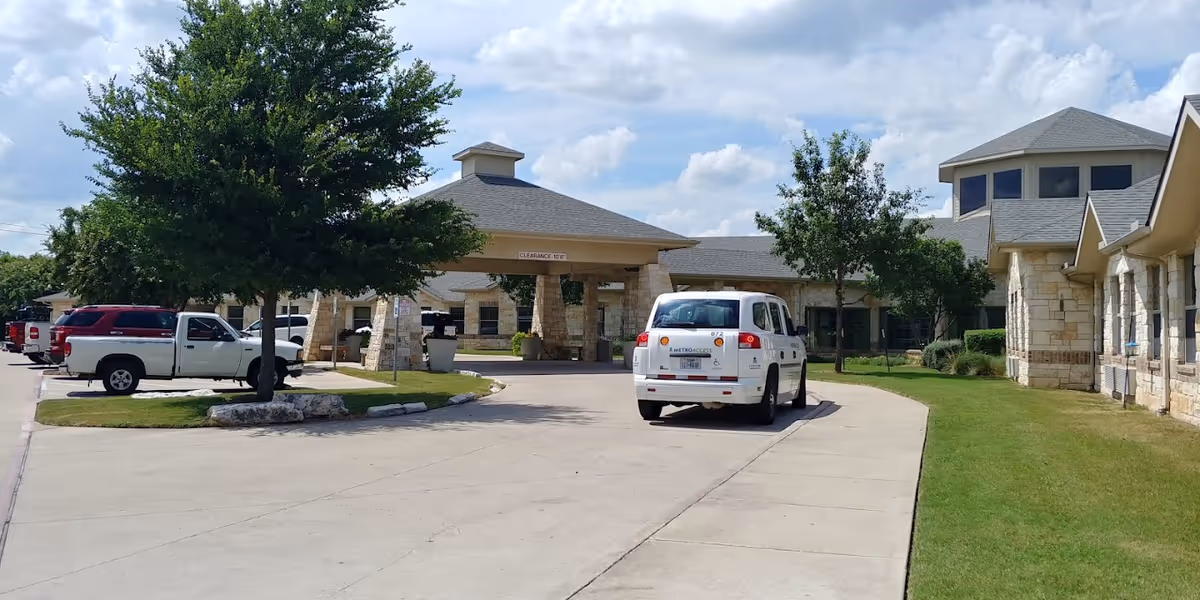 Driveway and covered entrance of a nursing and rehabilitation center with parked vehicles and stone-clad buildings.