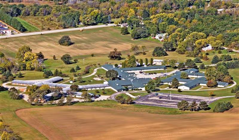 Aerial view of a low-rise senior living campus with multiple connected buildings, a parking lot, and surrounding fields and trees.