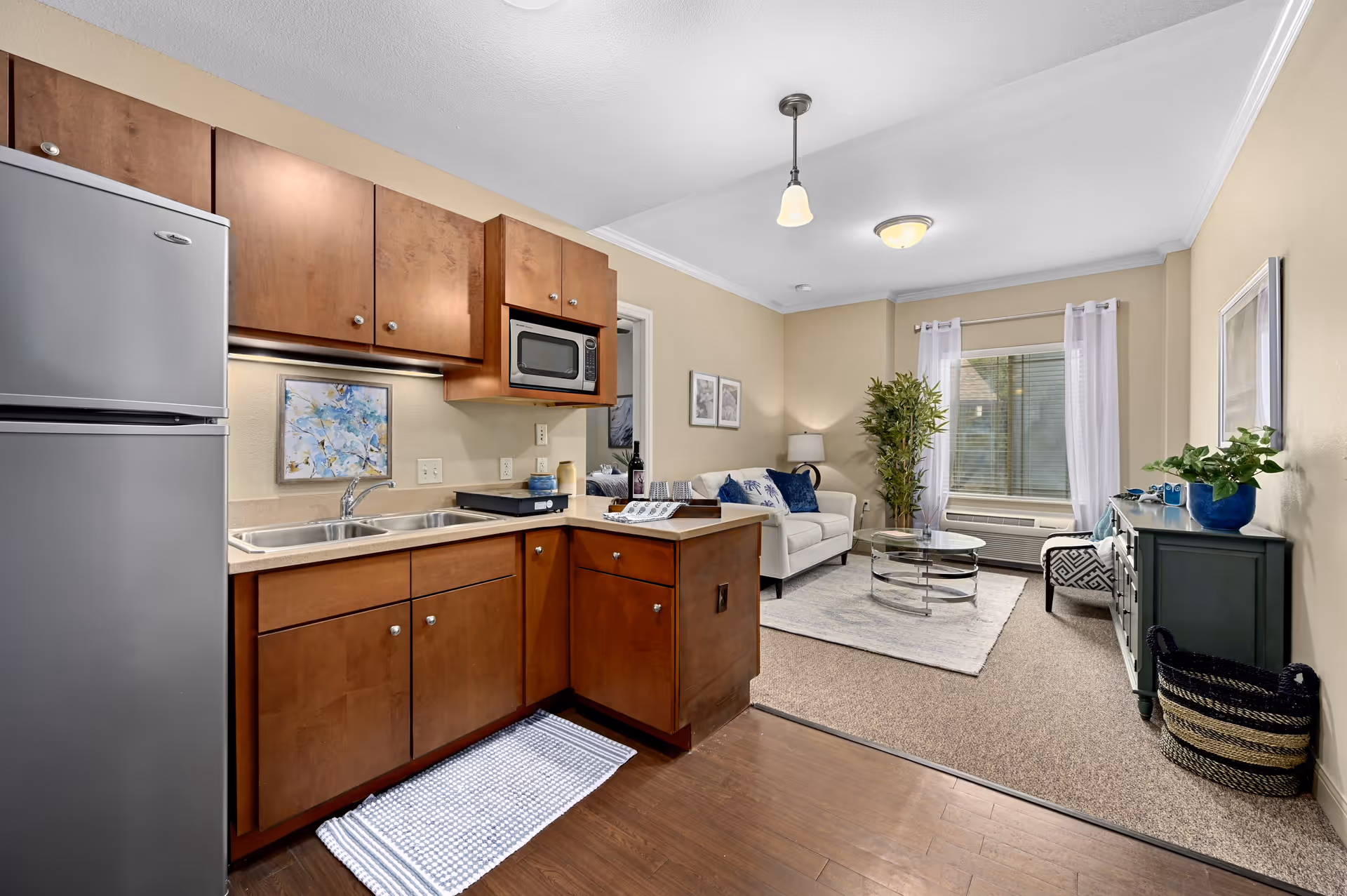 Interior view of a senior living facility apartment featuring a kitchen area with wooden cabinets, a stainless steel refrigerator, a double sink, and a microwave. Adjacent to the kitchen is a living room with a white sofa, glass coffee table, green sideboard, potted plants, and a window with white curtains allowing natural light in.