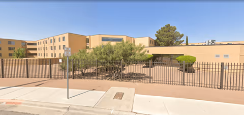 Exterior view of a beige senior living facility behind a metal fence with desert landscaping.