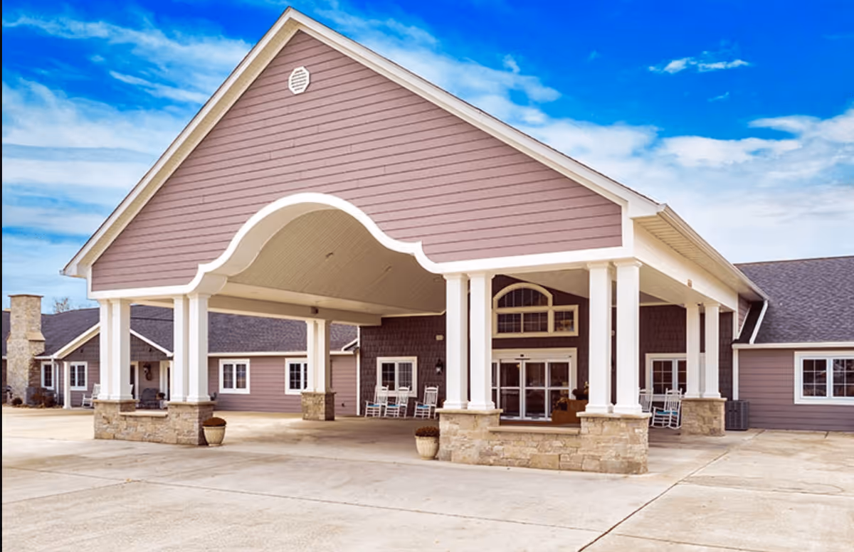 Front entrance canopy of a single-story senior living facility with white columns, rocking chairs, and a wide paved drive under a blue sky.