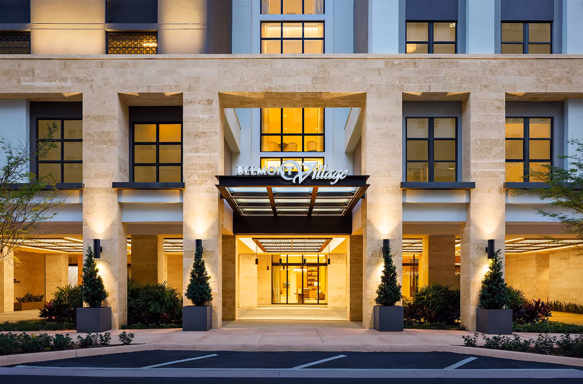 Front exterior view of Belmont Village Senior Living Coral Gables building entrance illuminated with warm lights, featuring large stone pillars, glass canopy, and potted plants along the walkway.
