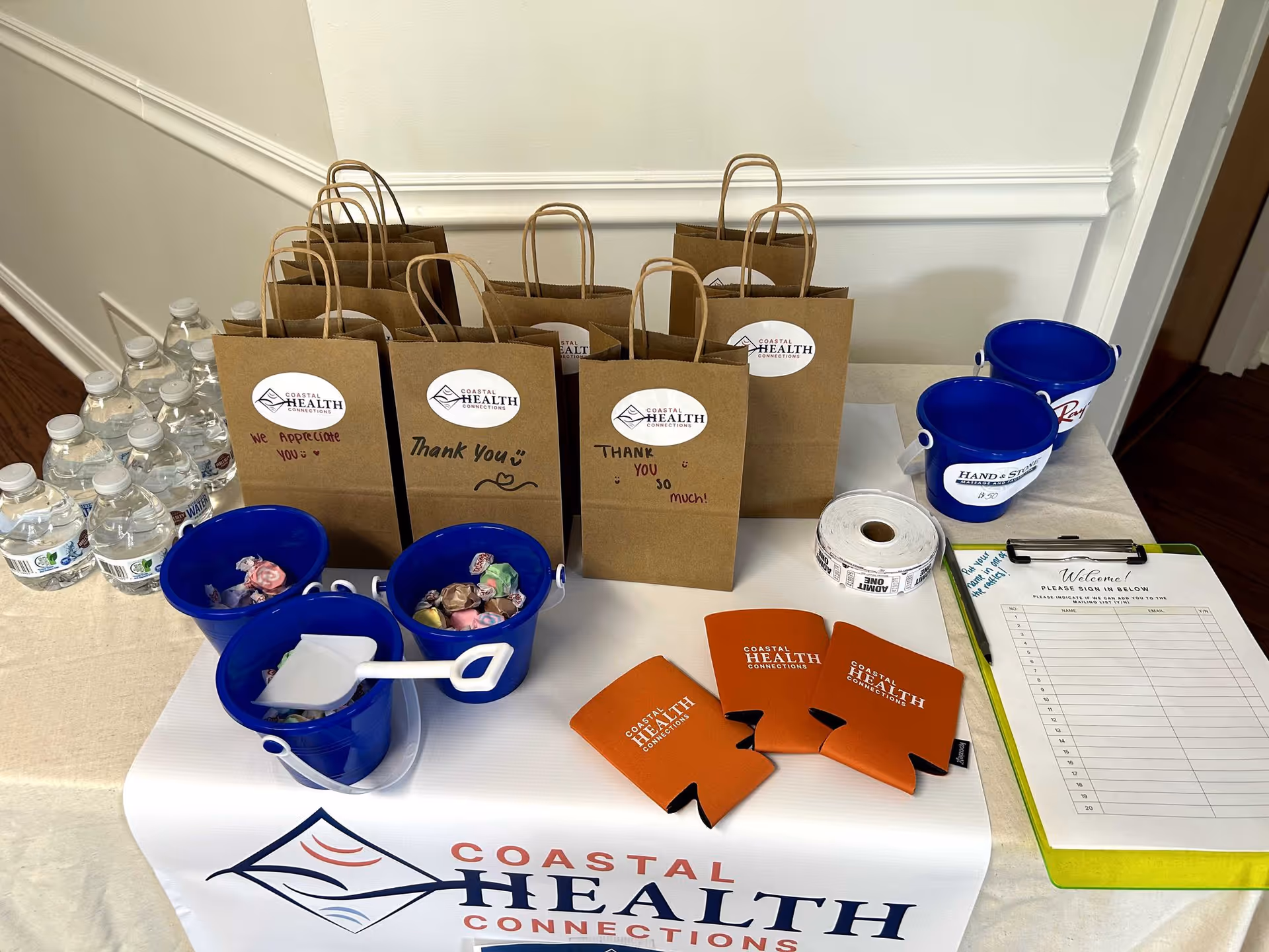 Table with Coastal Health Connections bags, blue bowls of candy, water bottles, and a sign-in clipboard.