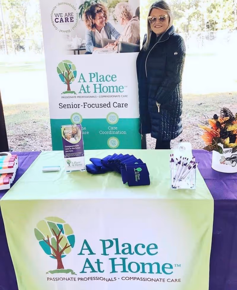 Outdoor promotional booth for A Place At Home The Woodlands with a staff member present.