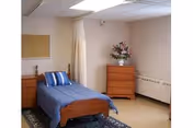 A furnished bedroom in a senior care home featuring a single bed, dresser, and flowers.