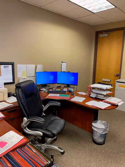 Office workspace with a desk, dual monitors, paperwork, and an empty chair.