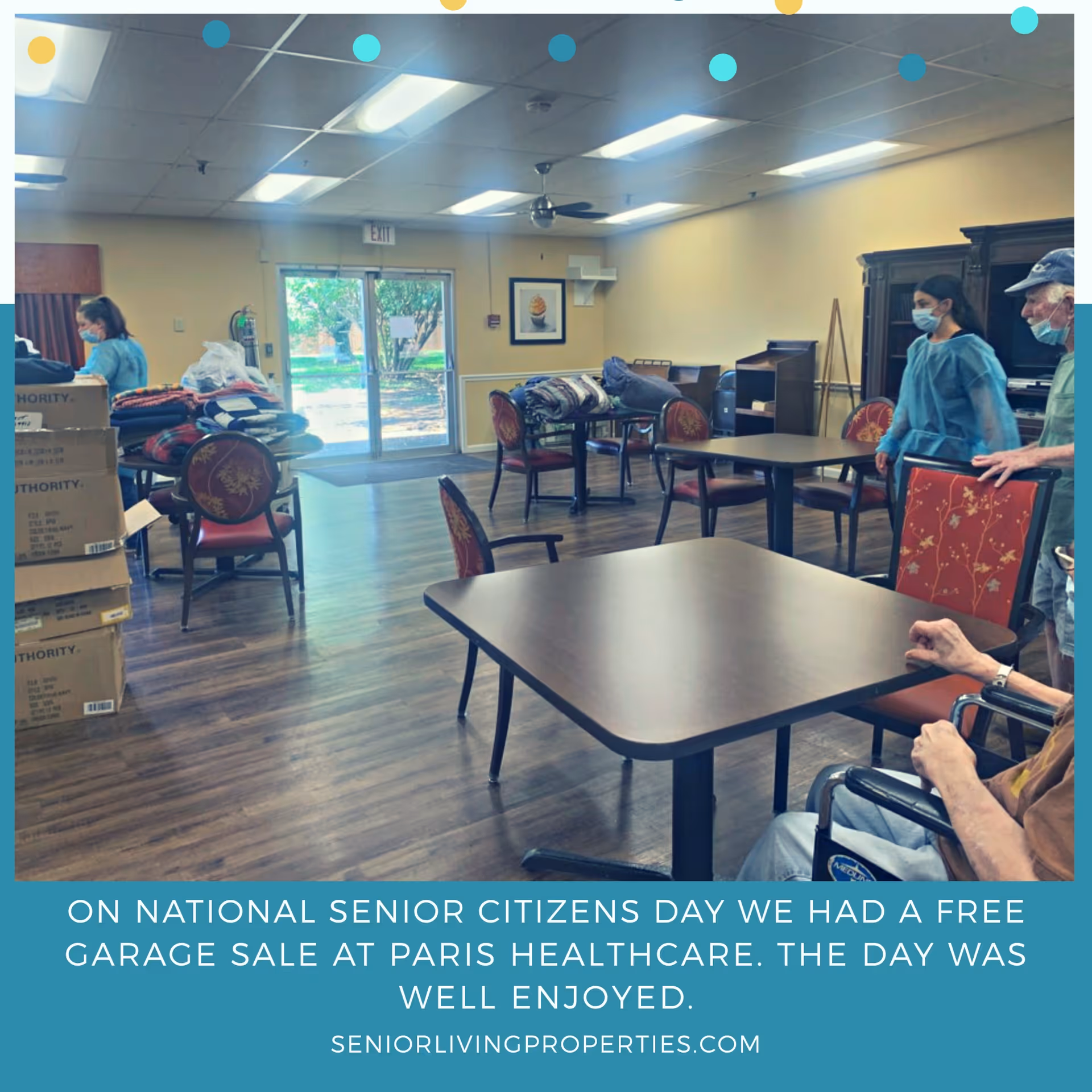Interior view of a common area in a senior living facility with tables and chairs arranged around the room. Several people, including staff wearing masks and seniors, are present. Boxes and folded blankets are stacked on some tables. Large windows and a glass door provide natural light and a view of the outdoors.