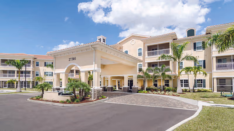 Exterior view of a beige senior living building with a covered entrance and palm trees.