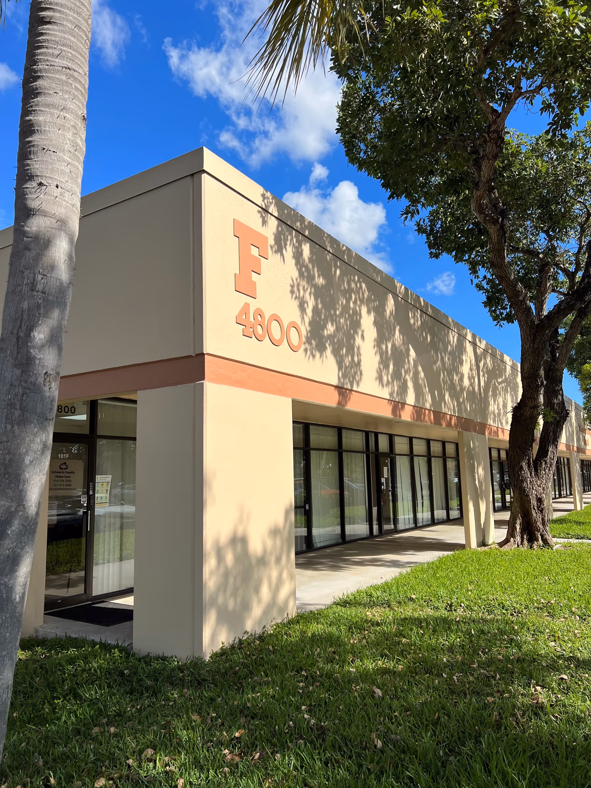 Beige building front with glass storefronts, a walkway, and palm trees under blue sky.