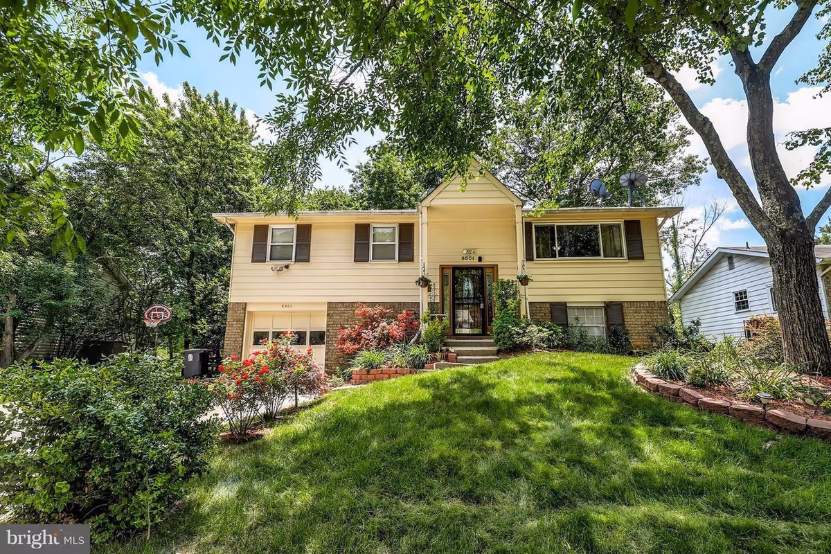 Two-story beige house with brown shutters, lush front yard, and trees framing the entrance.
