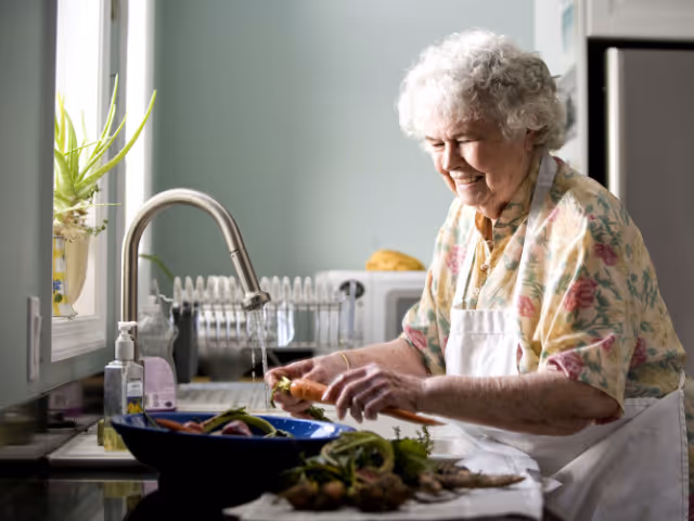 Elderly woman in an apron chops vegetables at a kitchen sink.