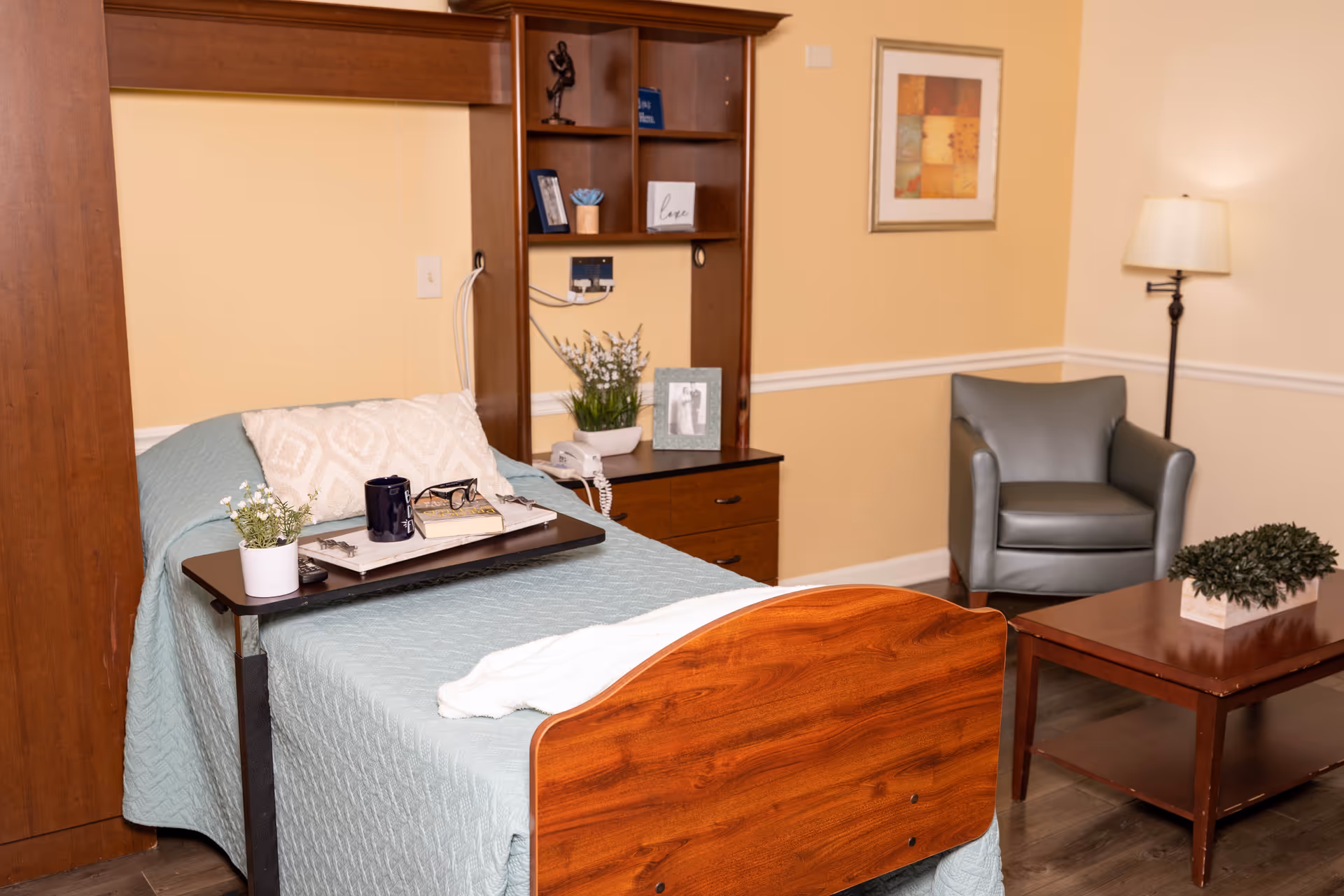 Senior living bedroom featuring a bed, desk, chair, plants, and warm beige walls.