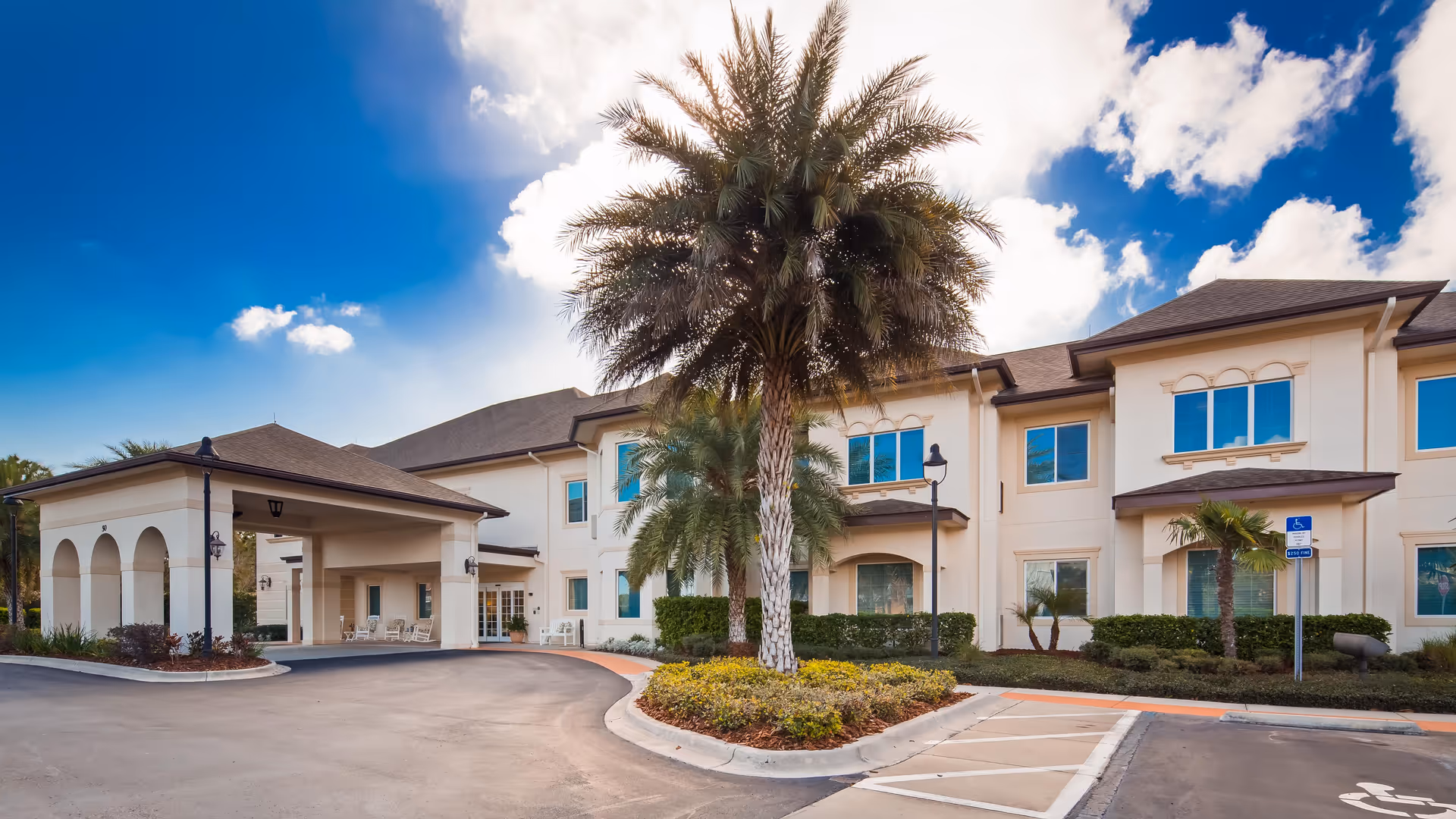 Exterior view of The Windsor of Palm Coast senior living facility showing a two-story building with a covered entrance, palm trees, landscaped bushes, and a clear blue sky with some clouds.