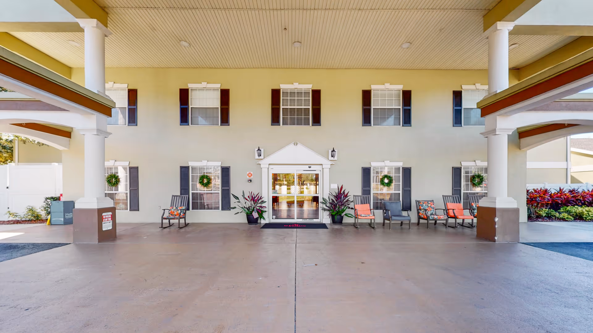 Covered entrance area of a senior living facility with a wide driveway, several chairs arranged along the wall, potted plants, and a double glass door entrance. The building facade has multiple windows with dark shutters and decorative wreaths.