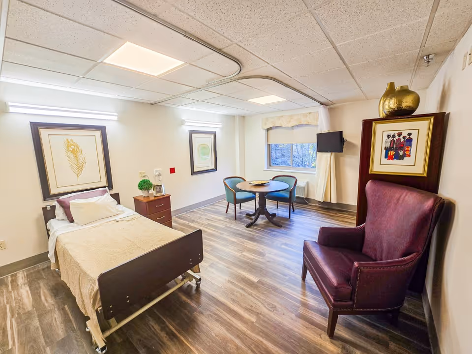 A furnished senior living bedroom with a bed, seating area, artwork, and window.