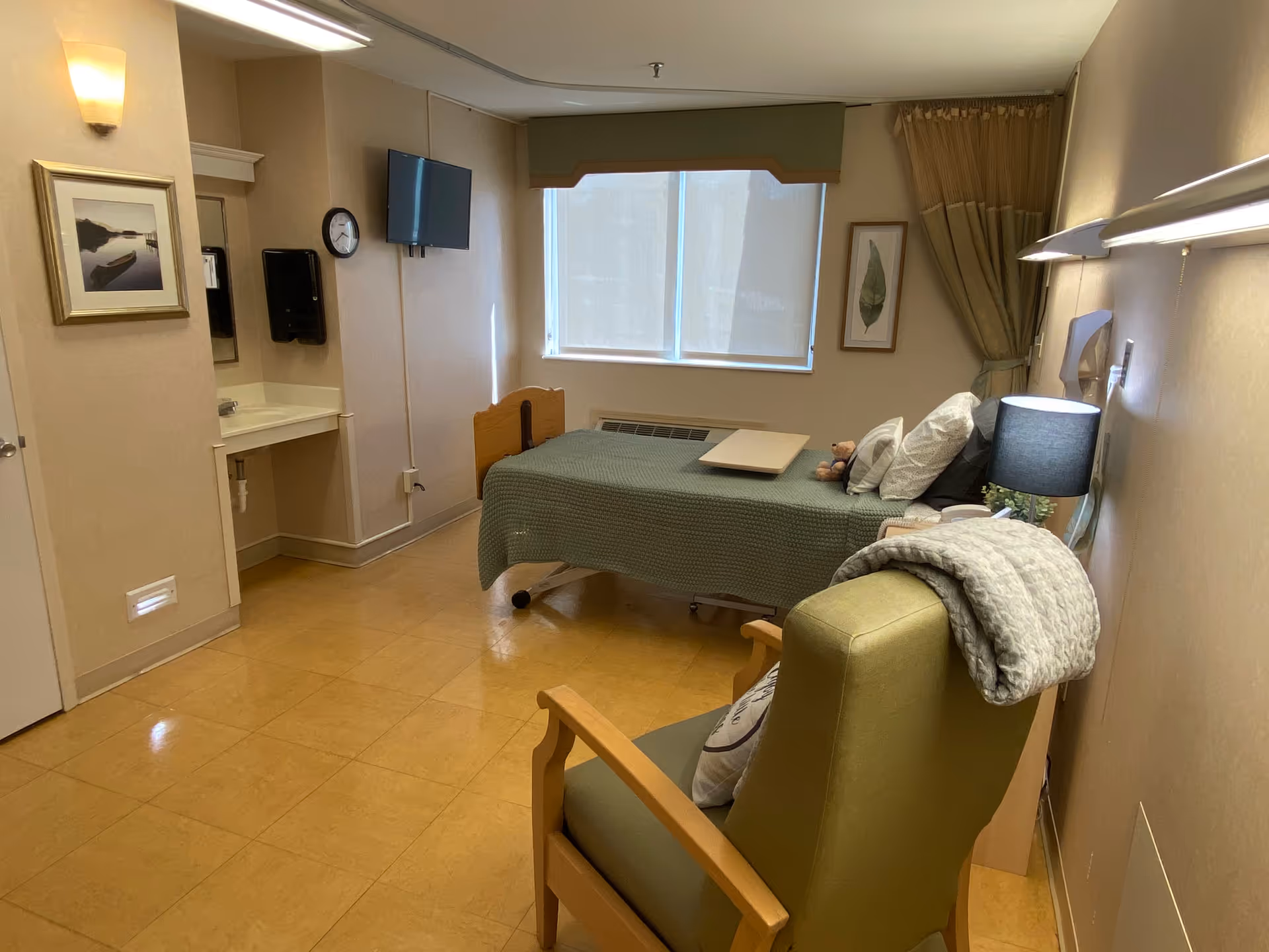 A hospital-style single-occupancy bedroom with a bed, chair, window, and wall decor.