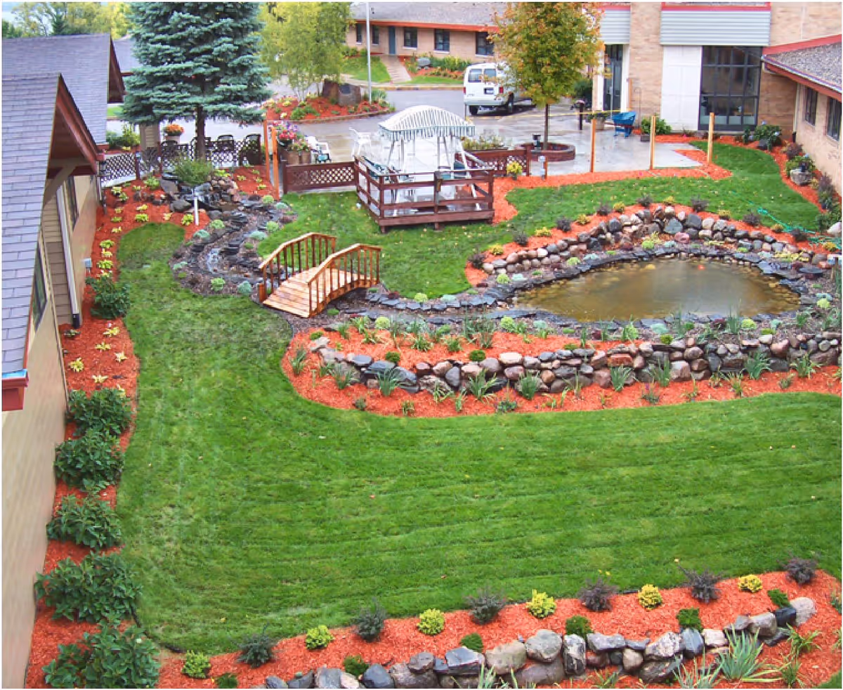 A landscaped outdoor courtyard with a pond, bridge, gazebo, and colorful garden in a care center.