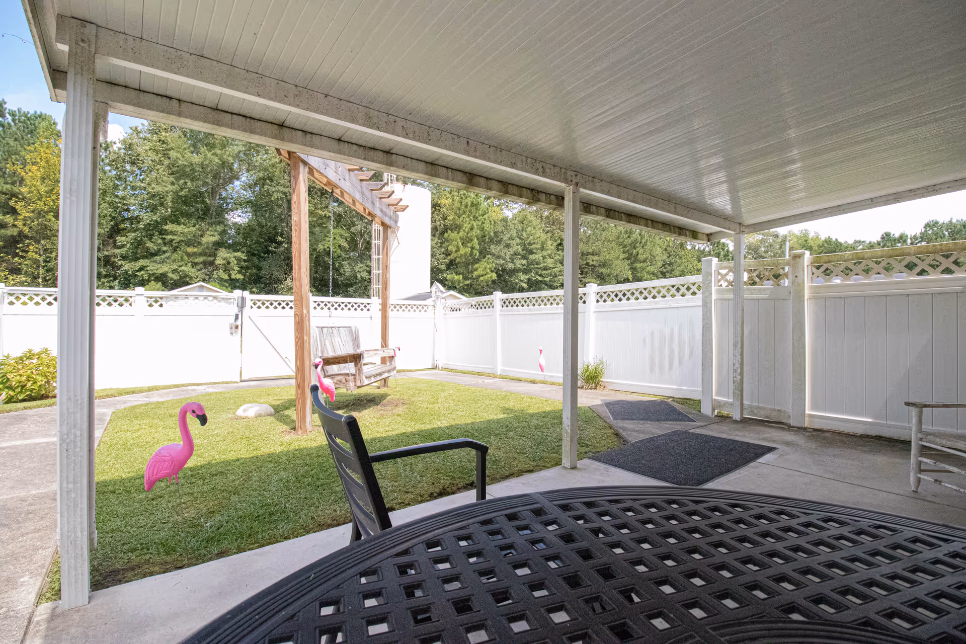 Covered patio with a table and chair overlooking a small fenced yard with a wooden swing and pink flamingo lawn ornaments.