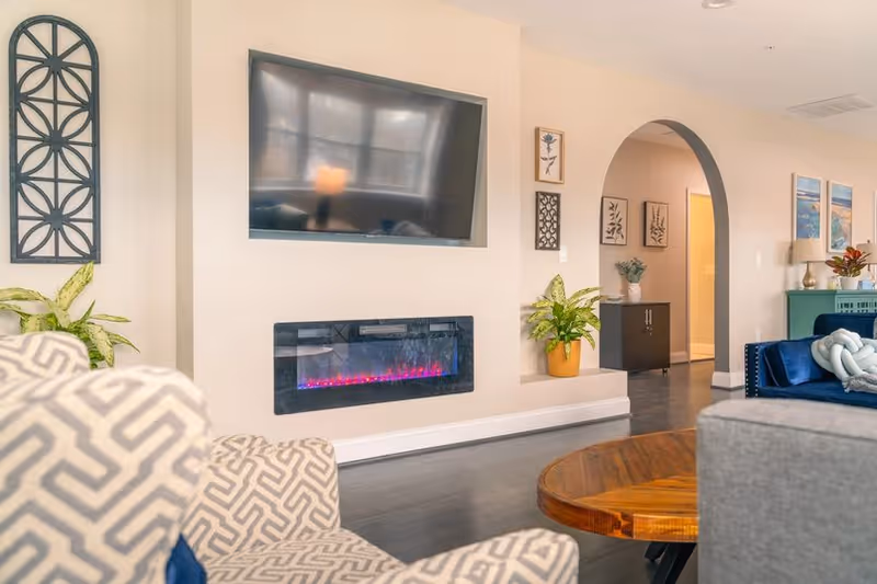 Modern senior living living room with TV, electric fireplace, plants, archway, and comfy seating.