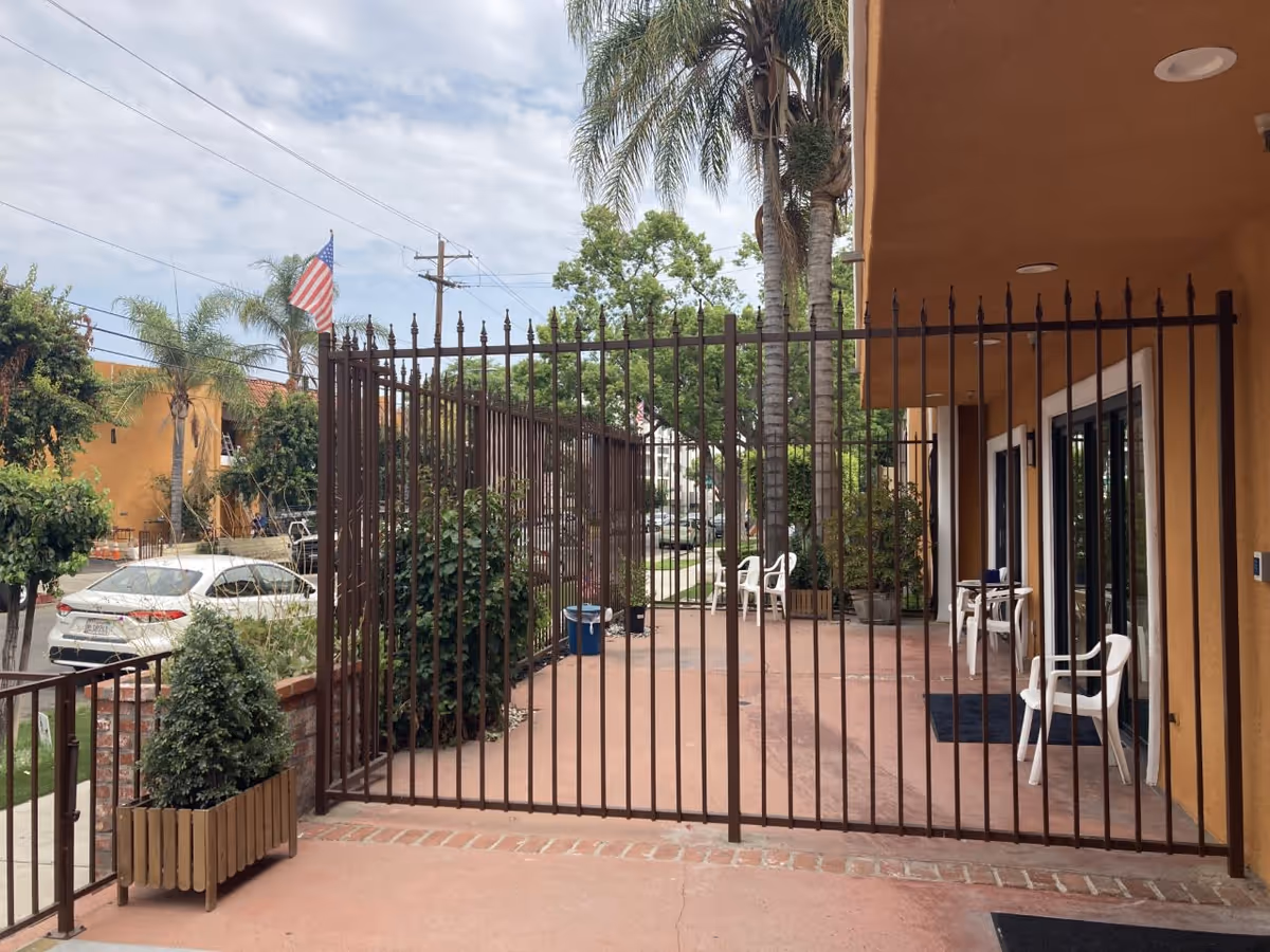 Outdoor patio area enclosed by a tall metal fence with vertical bars. The patio has a reddish concrete floor and is adjacent to a building with a brown exterior. There are several white plastic chairs and small tables on the patio. Palm trees and other greenery are visible around the area, with a street and parked cars in the background under a partly cloudy sky.