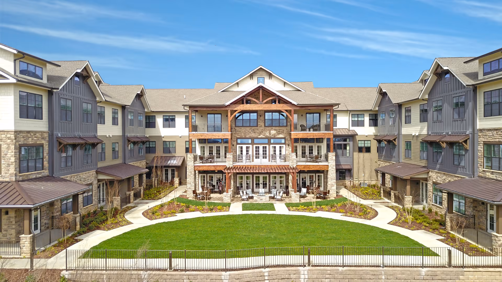Exterior view of a large, multi-story senior living facility with a central courtyard featuring a green lawn, paved walkways, and outdoor seating areas. The building has a combination of stone and siding with multiple windows and balconies under a clear blue sky.