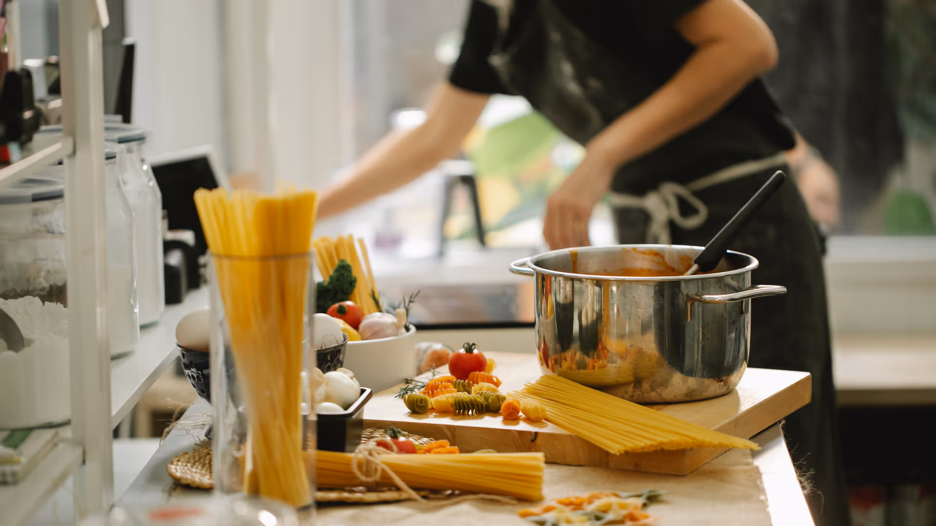 A person cooks in a kitchen with a large pot, pasta, and vegetables.