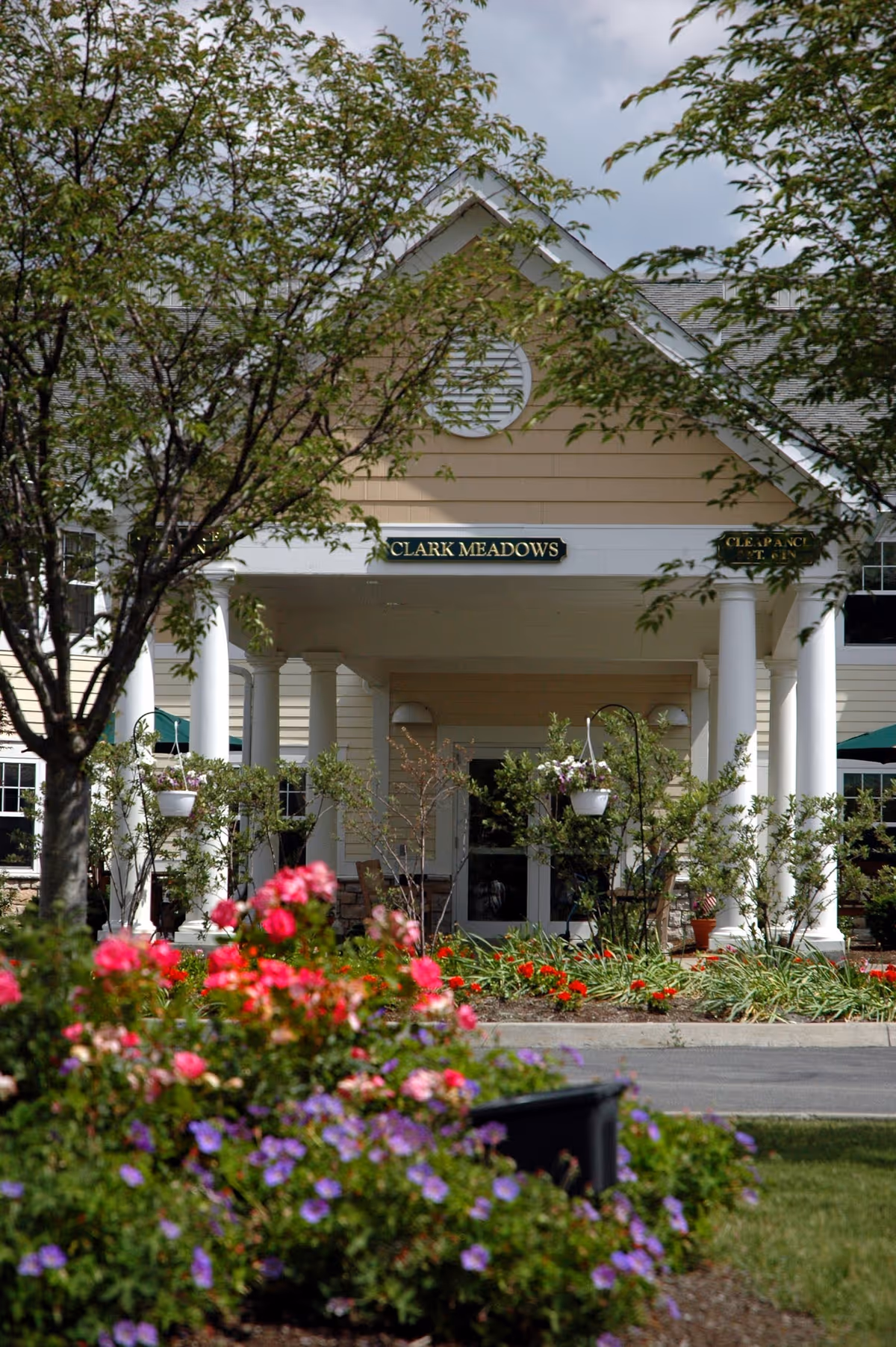 Entrance of Clark Meadows facility with a covered porch supported by white columns, surrounded by trees and colorful flower beds in the foreground.