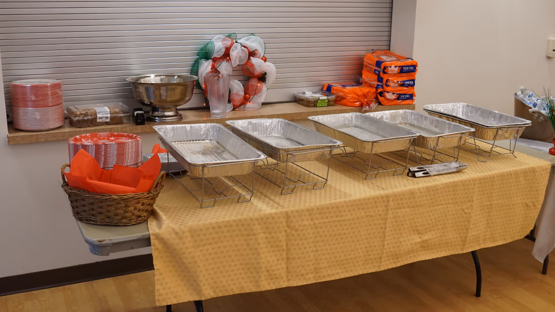 A buffet table set up with empty aluminum food trays on wire stands, a basket with red napkins, stacks of disposable plates, plastic cups, packaged food items, and a festive wreath on a counter behind the table.