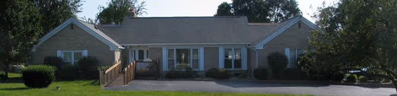Single-story senior living facility front exterior with ramp, shrubs, and parking area.