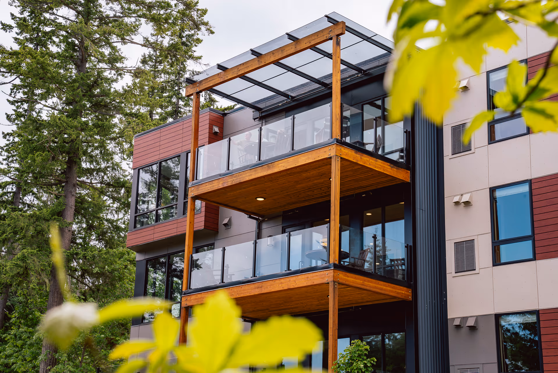 Modern multi-story senior living building with wooden balconies and glass railings among trees.