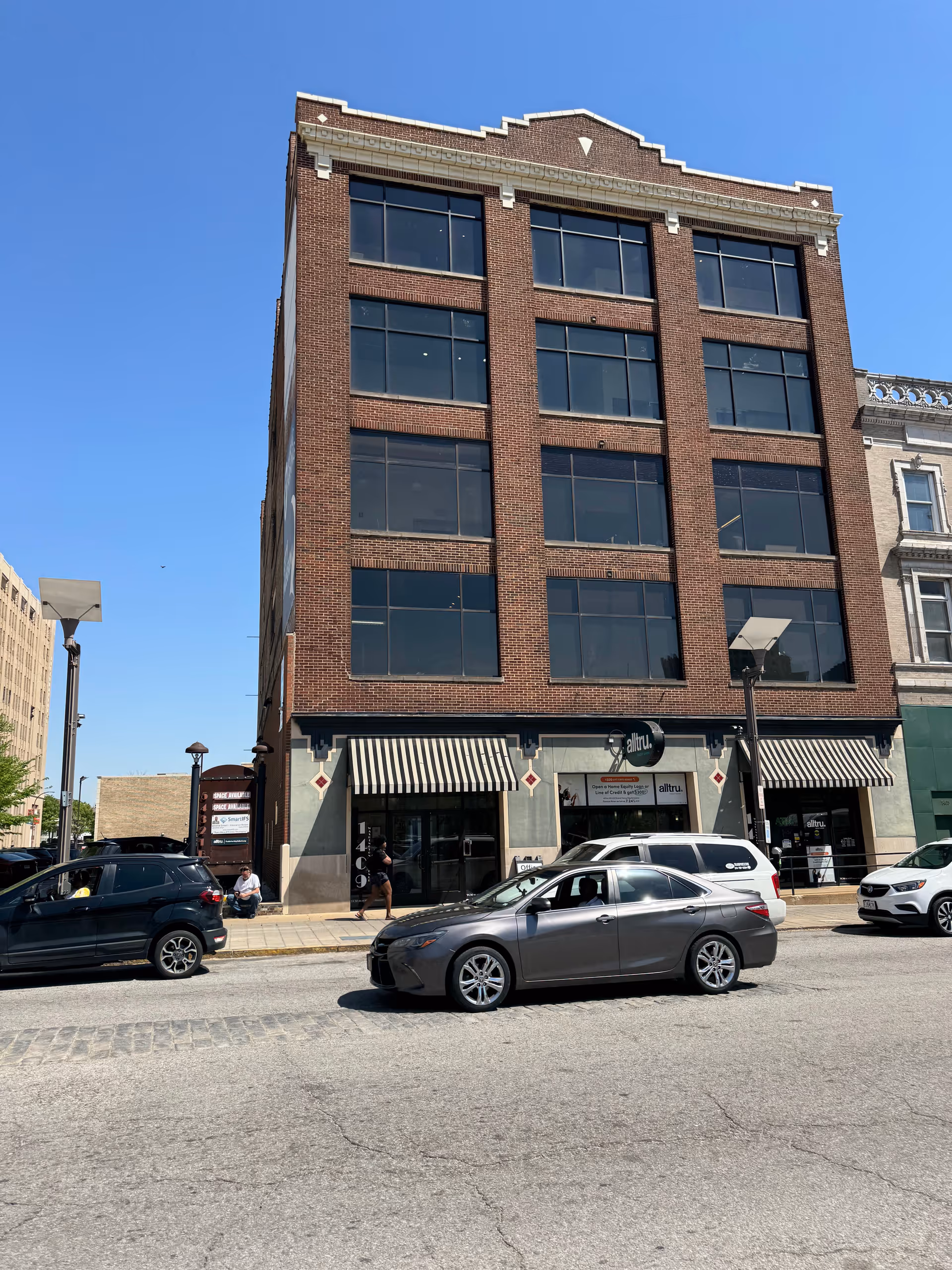 A tall brick building with large windows on a city street, storefronts at the ground level.