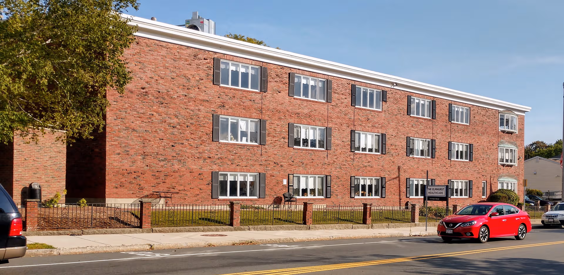 Three-story red brick senior living building with The Elmhurst HealthCare sign and parked cars.