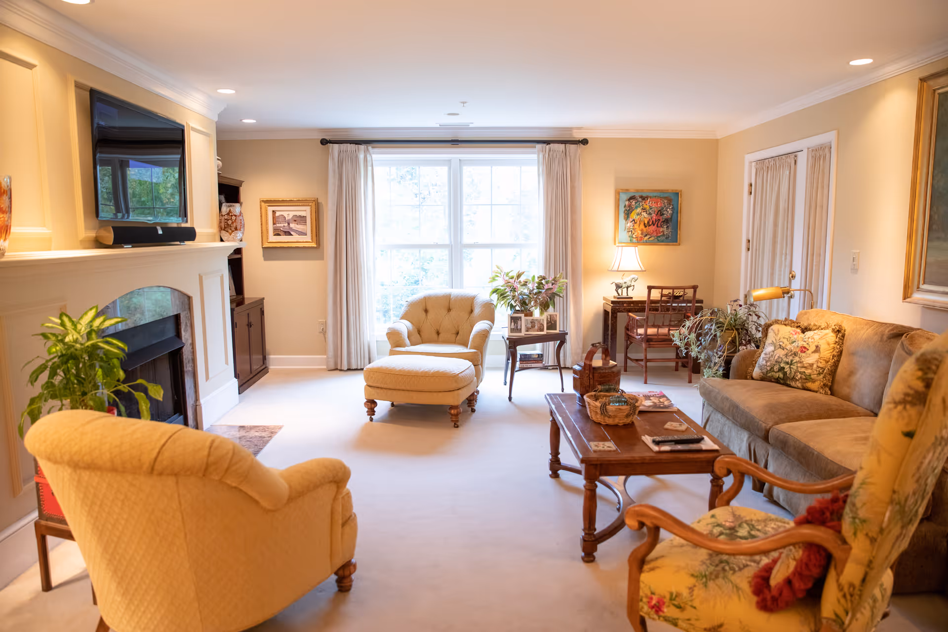 A cozy living room with beige walls and carpet, featuring a fireplace with a mounted TV above it, several upholstered chairs including a floral armchair and a beige armchair with an ottoman, a wooden coffee table with decorative items, a sofa with floral cushions, a side table with plants and framed photos, a small desk with a chair and lamp, and large windows with curtains letting in natural light.