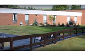 Single-story red brick nursing facility with windows, green lawn, and a wooden railing along walkway.