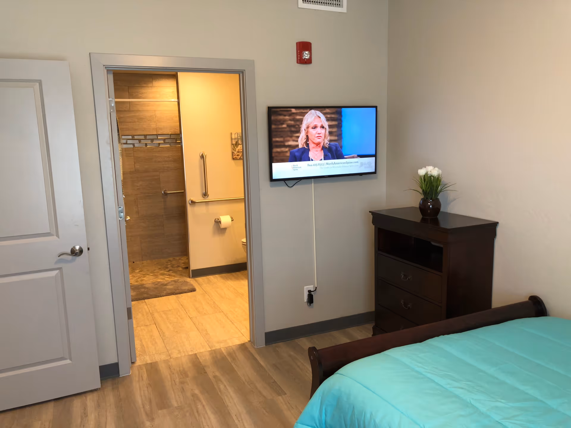 A bedroom with a teal bedspread, a dark wooden dresser with a vase of white flowers, and a wall-mounted TV showing a woman speaking. An open door reveals a bathroom with beige tiles, a grab bar, and a toilet paper holder.