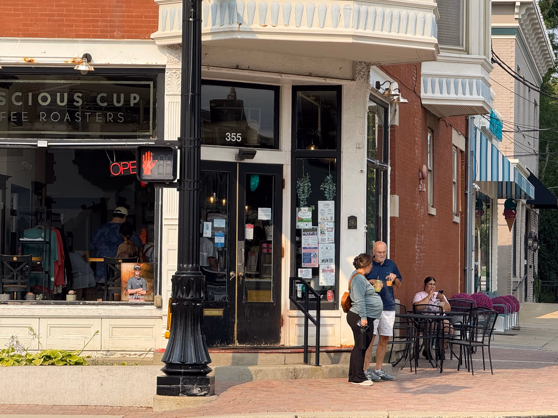 Busy street storefront with outdoor dining tables; two people chat near an entrance while passersby watch.