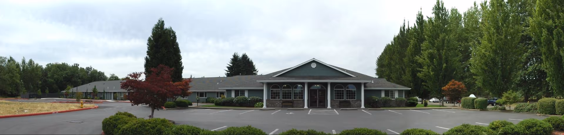 Panoramic view of a single-story memory care building with a central covered entrance, parking lot, and surrounding trees and landscaping.