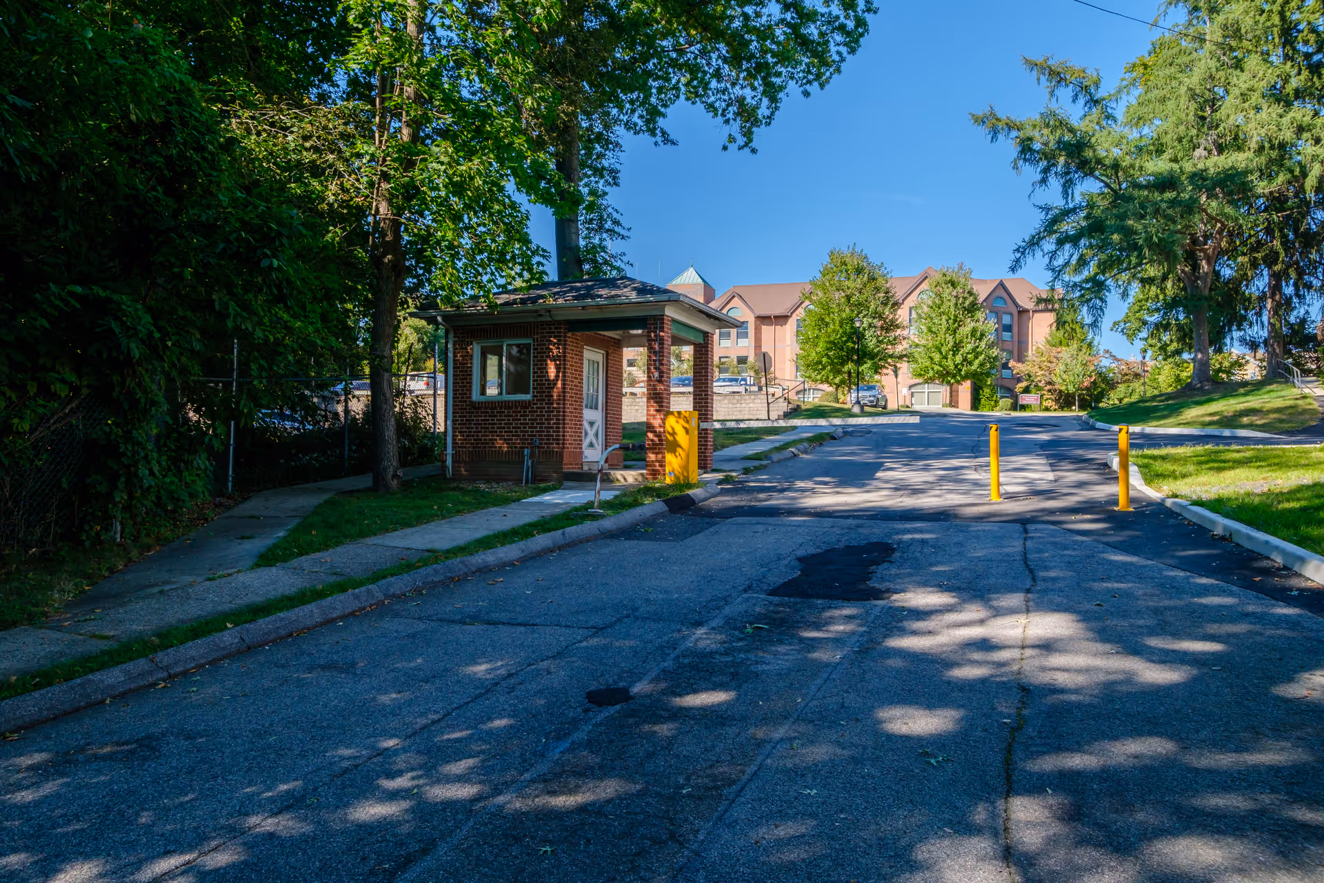 Driveway entrance with a small brick guardhouse and a multi-story assisted living building in the background framed by trees.