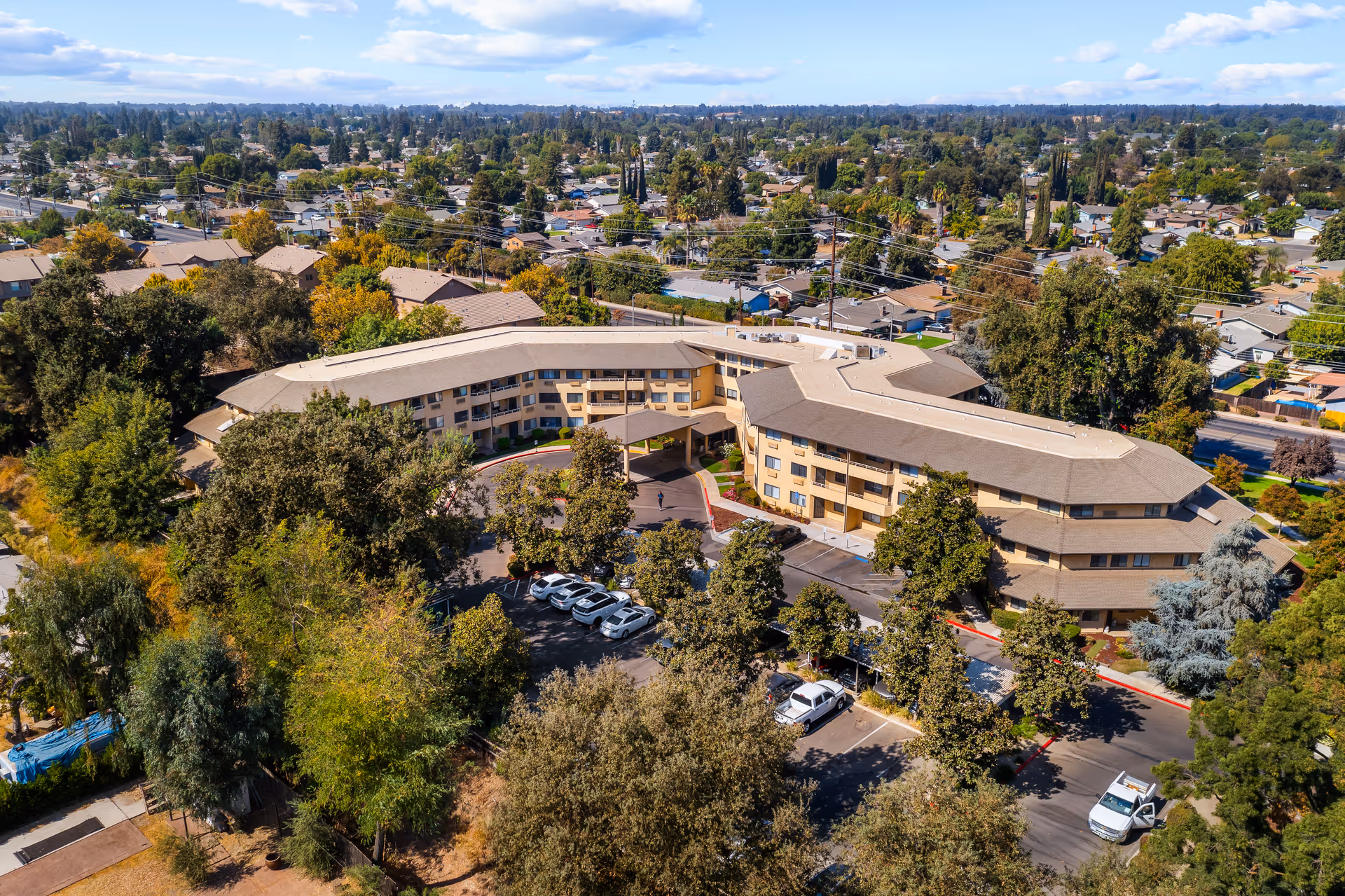 Aerial view of Walnut Park - A Provincial Senior Living Community, showing a large, three-story building with a beige exterior surrounded by trees and parking spaces. The building is situated in a suburban area with many houses and greenery visible in the background under a partly cloudy sky.