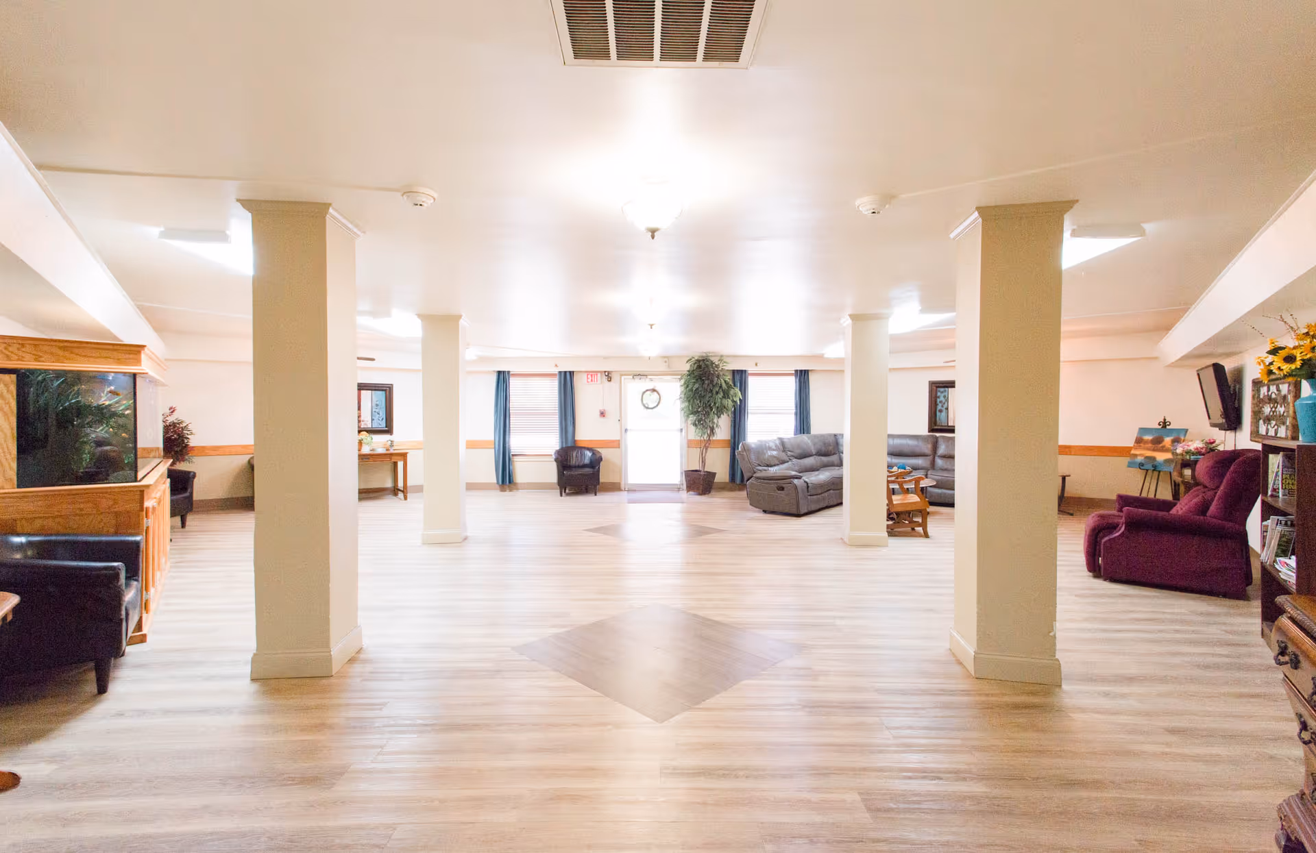 A spacious senior living facility common area with light wood flooring, cream-colored walls, and several support columns. The room is furnished with various seating options including leather sofas, armchairs, and a rocking chair. There is a large fish tank on the left side, a potted plant near the center back door, and a bookshelf with flowers and decorations on the right. Windows with blue curtains allow natural light into the room.